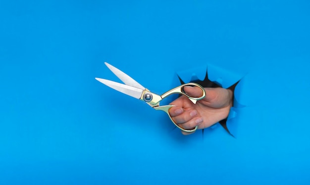 Female hand torn a hole in blue paper wall and holding gold sewing scissors cut the price or taxes concept cutting and sewing lessons