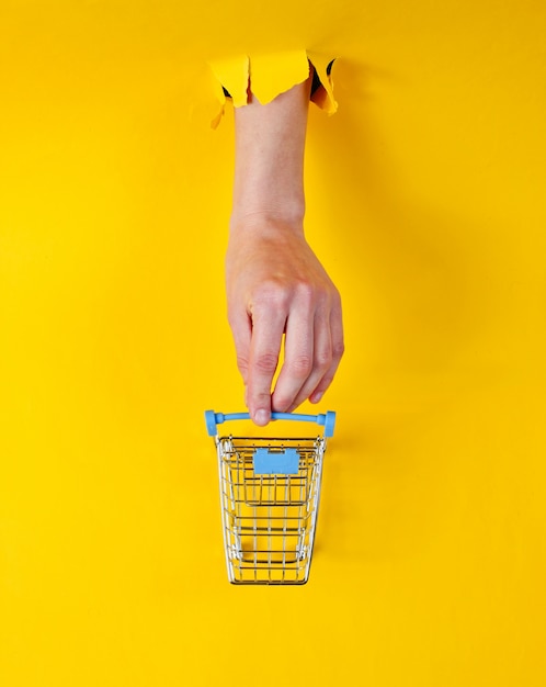 Female hand touches a mini shopping trolley through torn yellow paper. Minimalistic shopping concept