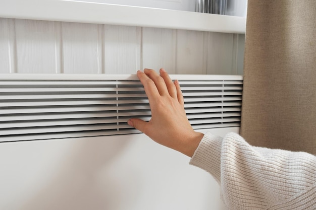 Female Hand warming over electric Heater Frozen Woman wearing a warm woolen Sweater freezing for winter Cold Discomfort spending time at Home Girl's Hands on modern white Radiator Heating problems