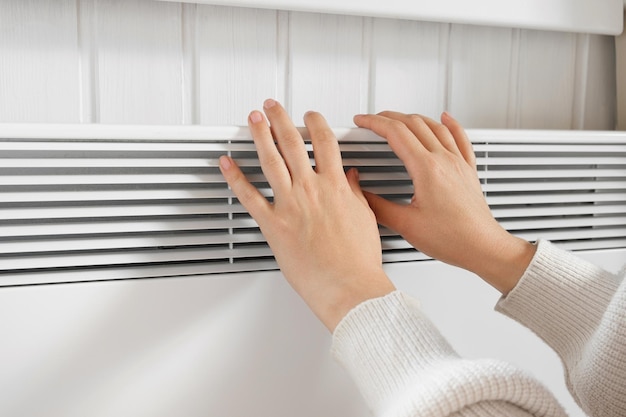 Female Hand warming over electric Heater Frozen Woman wearing a warm woolen Sweater freezing for winter Cold Discomfort spending time at Home Girl's Hands on modern white Radiator Heating problems