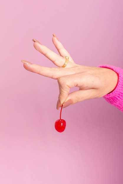 Female hand wearing pink sweater and holding a cherry. Light pink background.