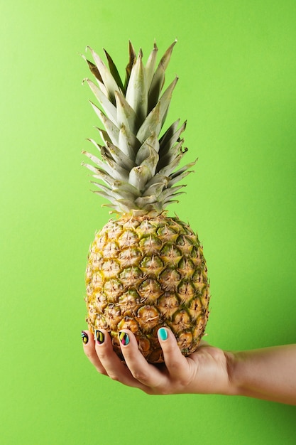 Female hand with colorful manicure holding pineapple on green background