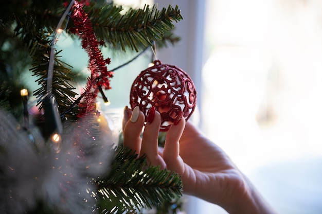ライトが付いているクリスマスツリーからぶら下がっている光沢のある赤い休日の安物の宝石を保持している休日のマニキュアと女性の手