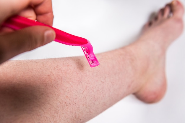 Female hand with pink razor shaves her legs on gray wall.