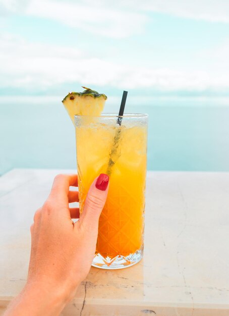 Photo female hand with a red manicure holds a tropical yelloworange cocktail on the background of the sea