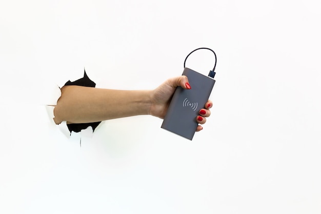 A female hand with a red manicure through torn white paper holds a power bank