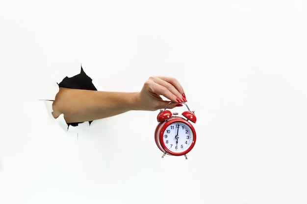 Photo a female hand with red manicure through torn white paper holds a red alarm clock. hand through torn white paper. hand holding a red alarm clock