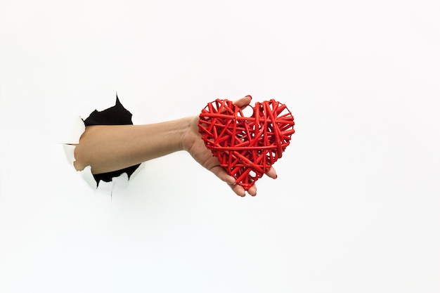 Foto una mano femminile con una manicure rossa attraverso carta bianca strappata tiene un cuore rosso