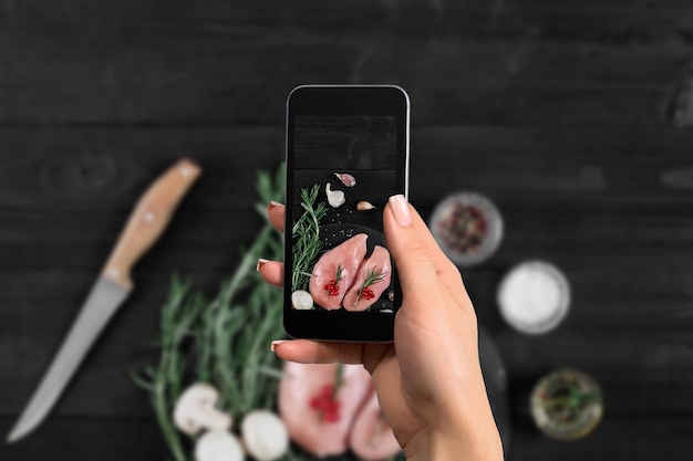 Женская рука со смартфоном делает фото сырой куриной грудки на черном фоне со свежими грибами ...