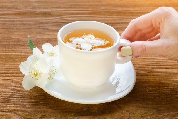 Female hand with white mug of tea and jasmine on a wooden table. Close up