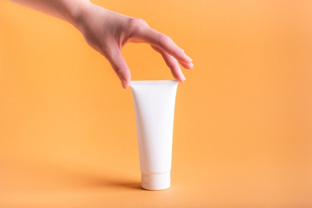 Female hand with white tube of body cream on orange background