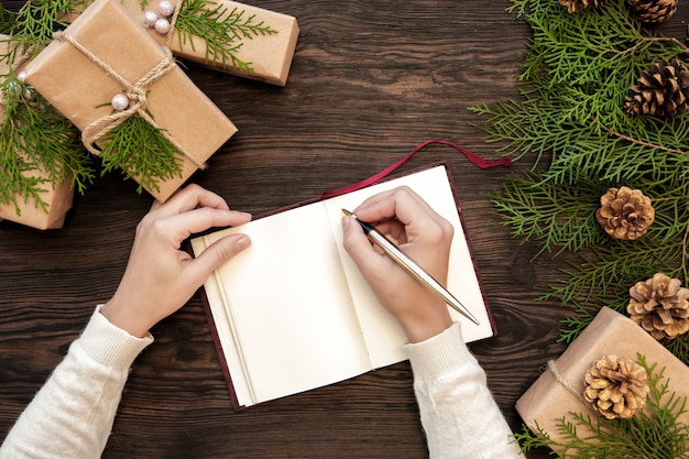 Female hand writes Christmas letter in notebook on dark boards with gifts and fir cones