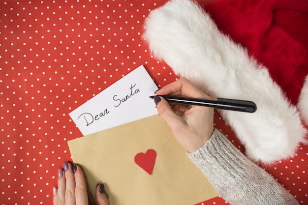 Female hand writes letter to Santa, envelope. Santa hat, red surface.