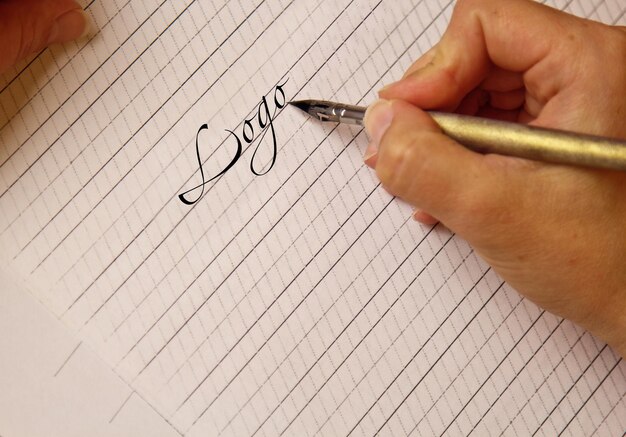女性の手はインクのペンで白い紙のシートに単語の文字を書き、机の上の縞模様の文房具を接写、上面図のスペル レッスンと書道演習テンプレート レイアウトの背景