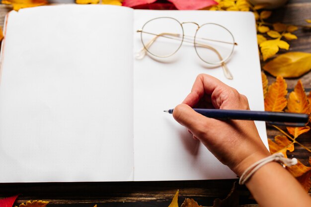 Foto mano femminile scrivendo in un diario di proiettile. la pagina in bianco del blocco note con le donne circonda i vetri sulla cima in uno spazio accogliente con le foglie arancio, gialle e rosse di autunno su una tavola di legno.