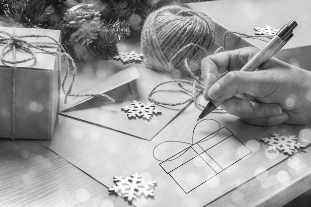 Foto mano femminile che scrive una lettera di natale per babbo natale, disegna una scatola regalo sul tavolo con una busta, regali di natale, abete e fiocchi di neve, stile ecologico, bokeh festivo, bianco e nero