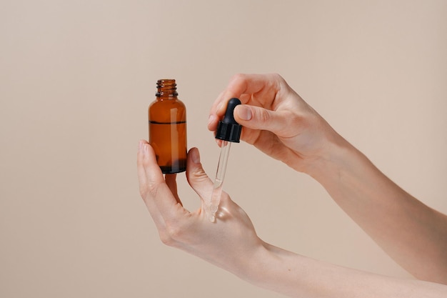 Female hands applying natural blank serum to skin from brown glass jar on beige isolated background Concept of beauty spa treatment and aesthetics