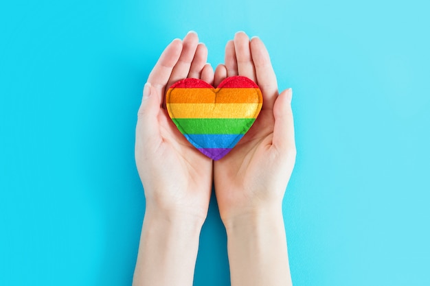 Female hands are holding a rainbow heart symbol of the LGBT community on a blue background, greeting card, background for poster, flyer, banner, copy space. LGBT day concept. LGBT background.