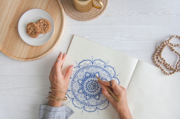 女性の手をクローズアップ描画装飾的な丸い花曼荼羅。趣味とリラクゼーション。木製トレイ上のコーヒーとクッキーのマグカップ。白色の背景。