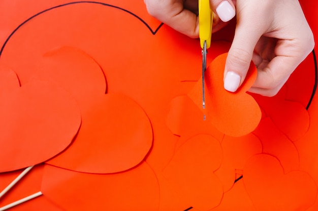 Photo female hands cut a red heart with scissors on a red paper background. preparation for the holiday.