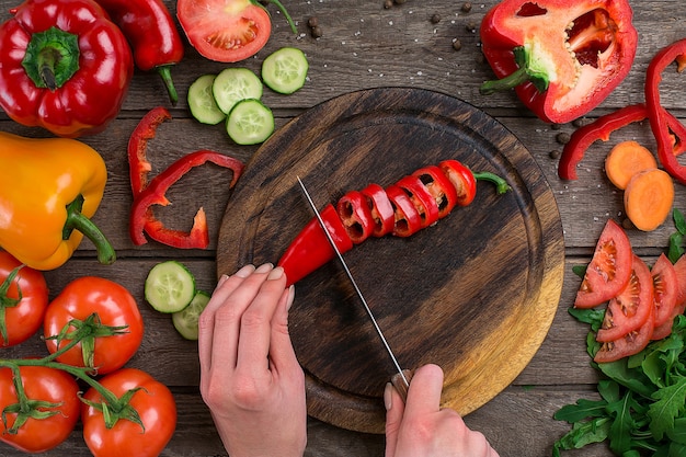 Female hands cutting hot peppers at table, top view. On the table vegetables and a wooden board