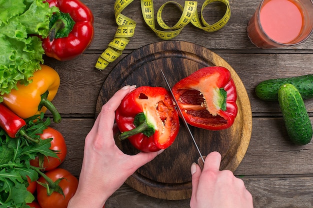 Female hands cutting pepper at table, top view. On the table leaves of lettuce, pepper, a glass of tomato juice, a wooden board and a knife