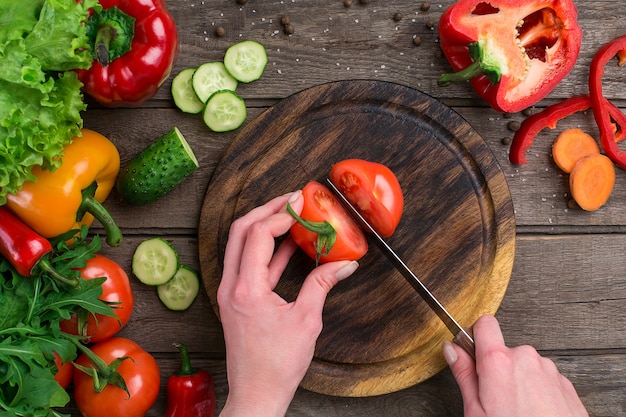 Female hands cutting tomato at table, top view. On the table vegetables and a wooden board