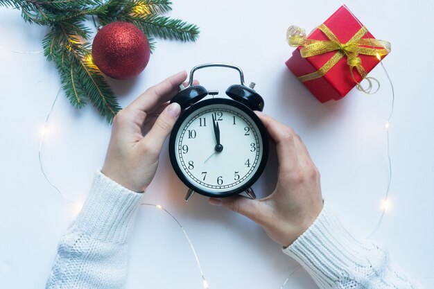 Female hands, dressed in a white sweater, holding a large black alarm clock showing twelve o'clock.