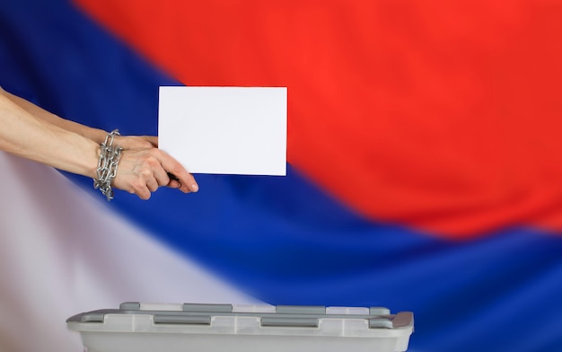 금속 사슬로 고정된 여성의 손은 투표함에 투표 용지를 던졌습니다. 백그라운드에서 러시아 국기입니다.