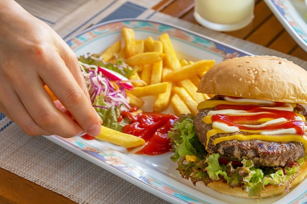 Female hands fingers holding and dripping fried French fries in tomato ketchup on a plate with hamburger