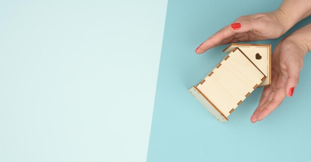 Female hands folded to each other over a wooden miniature model house on a blue background rea