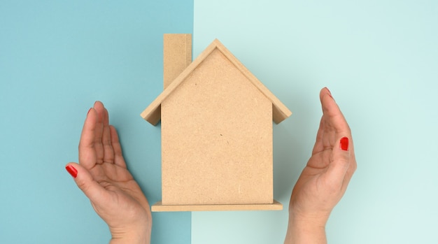 Female hands folded to each other over a wooden miniature model house on a blue background. Real estate insurance concept, environmental protection, family happiness