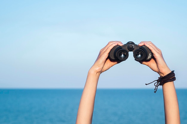 Female hands hold binoculars on the blue horizon sky and sea background