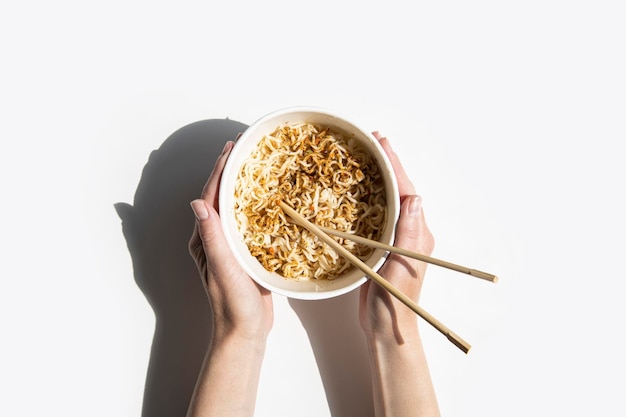 Female hands hold Chinese noodles in a cardboard bowl on a white background Top view fly lay