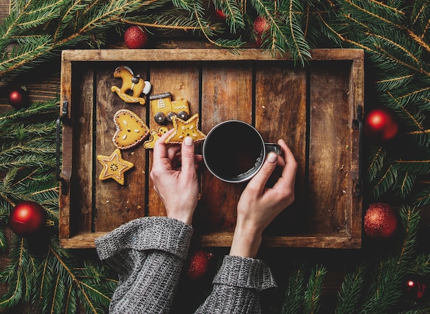 女性の手は、トレイのクッキーとクリスマスツリーの横にお茶を持っています