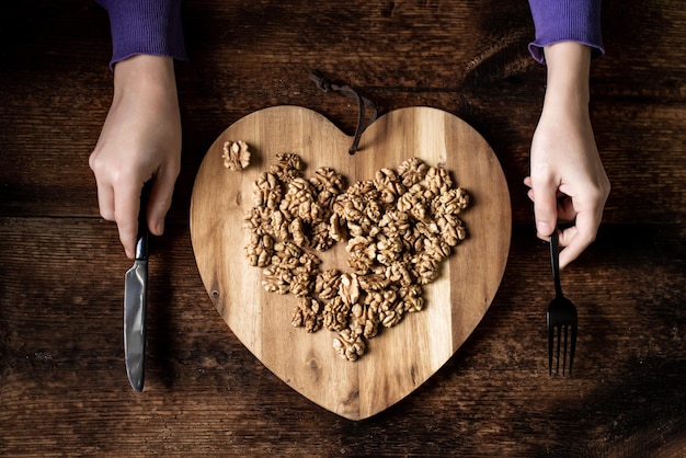 Female hands hold cutlery a knife and a fork in their hands peeled walnut on a wooden tray in the shape of a heart Dark wooden background Healthy eating Vegetarianism