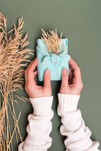 Female hands hold eco friendly furoshiki gift with ears of dry grass on green background