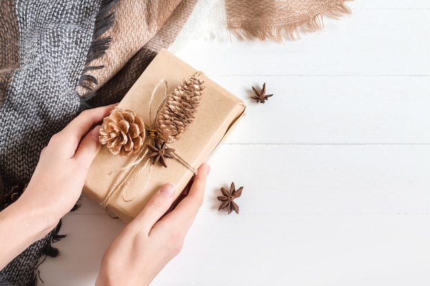 Female hands hold a gift box in craft paper with anise, dry cones, orange and cinnamon stick on a white wooden  in rustic style, copyspace, top view