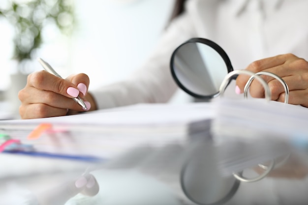 Female hands hold magnifying glass and pens next to documents