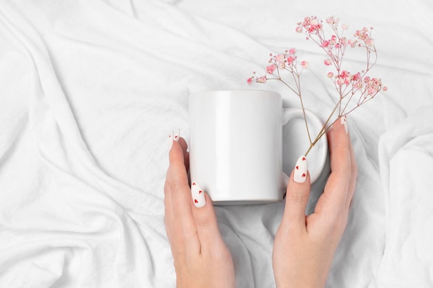 Female hands hold mock up white empty mug on a textile background cup for Valentine's Day manicure