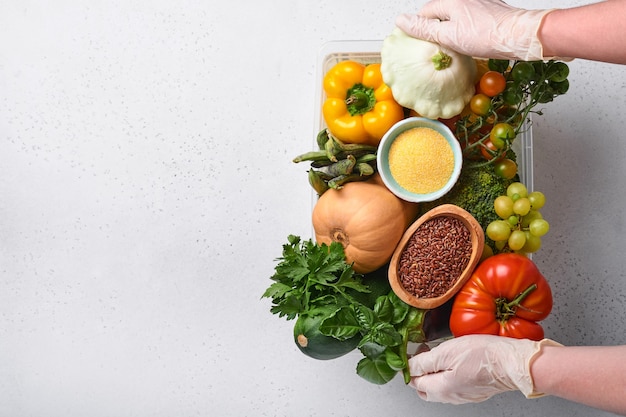 女性の手は、古い木製の背景に新鮮な野菜、果物、シリアル、種子の品揃えのプラスチックボックスを保持します。安全な宅配、食品調理、健康的なきれいな食品の背景とモックアップ。