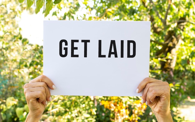 Female hands hold a sheet of white paper overhead. On the sheet the inscription GET LAID.