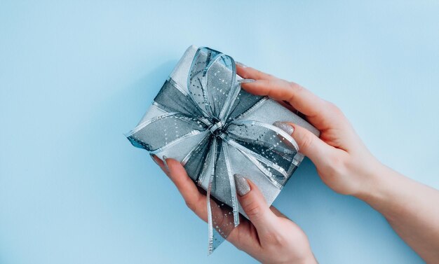 Female hands hold Silver gift with ribbon bow on blue background Holiday present christmas new year birthday valentine's day 8 march Flat lay Top view copy space