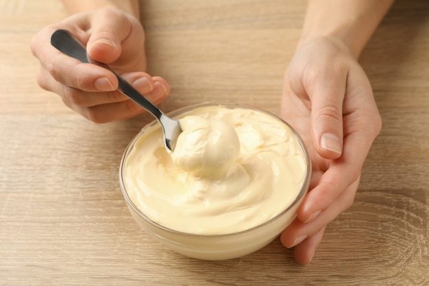 Female hands hold spoon and bowl with mayonnaise on wooden background