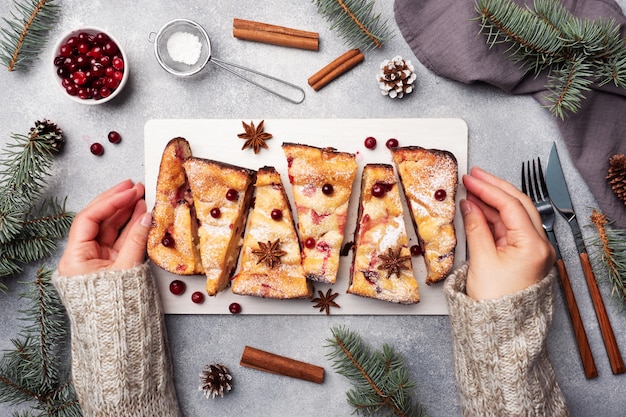 Female hands hold a stand with a Cottage cheese pie casserole with cranberries and spices sprinkled with powdered sugar. Gray concrete table.