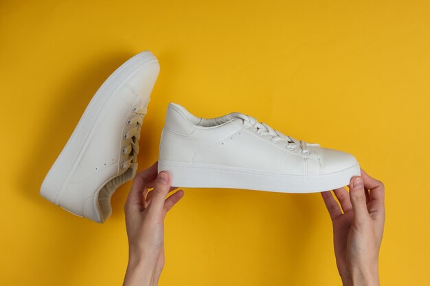 Female hands hold white stylish sneakers on a yellow studio paper