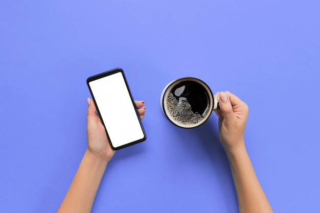 Mani femminili che tengono telefono cellulare nero con schermo bianco vuoto e tazza di caffè