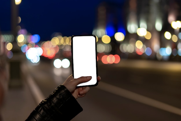 Foto mani femminili che tengono smartphone a schermo vuoto durante la notte di strada in città.