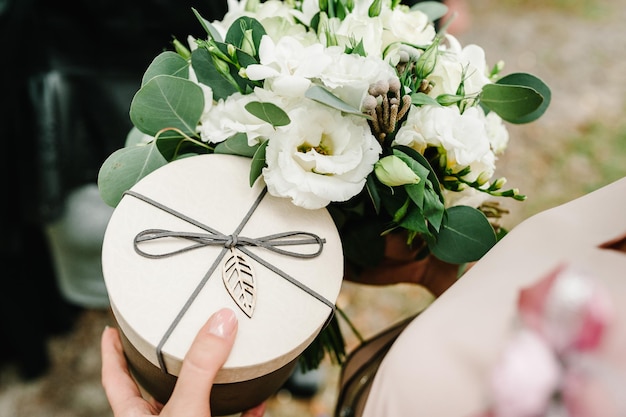 Female hands holding bouquet of flowers and gift round box with lid, flat lay. Summer stylish composition. Woman give brown box. Happy Mother's Day, Women's Day, Valentine's Day or Birthday.