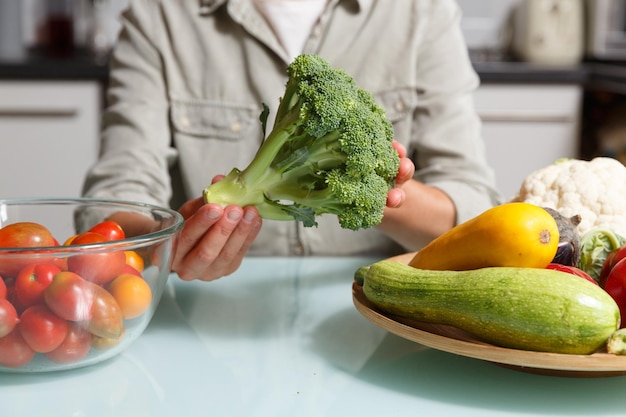 さまざまな野菜と一緒にキッチンの背景にブロッコリーを持つ女性の手健康食品調理のコンセプト